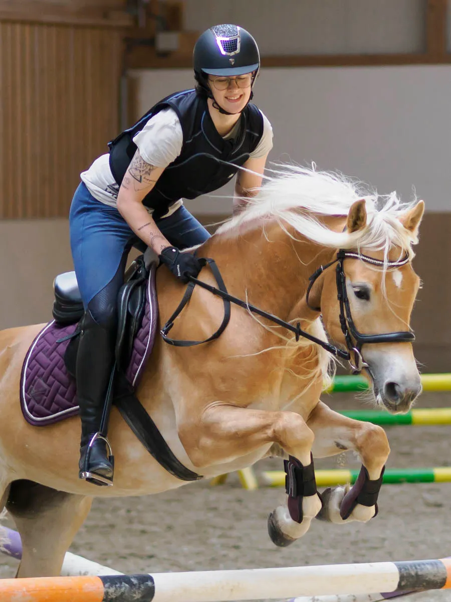 Lachende Reiterin mit Brille springt auf einem Haflinger mit wehender Mähne über ein Hindernis in der Reithalle