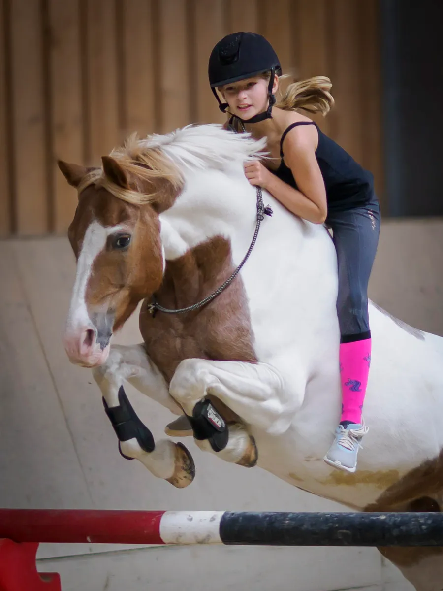 Mädchen springt ohne Sattel auf braun-weißem Schecken-Pony über ein Hindernis in der Reithalle