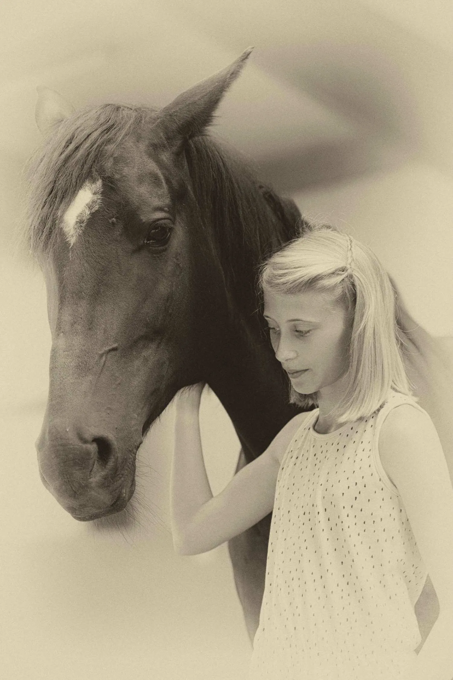 Blondes Mädchen steht mit gesenktem Blick an der Seite eines dunklen Pferdes mit Blesse, Sepia