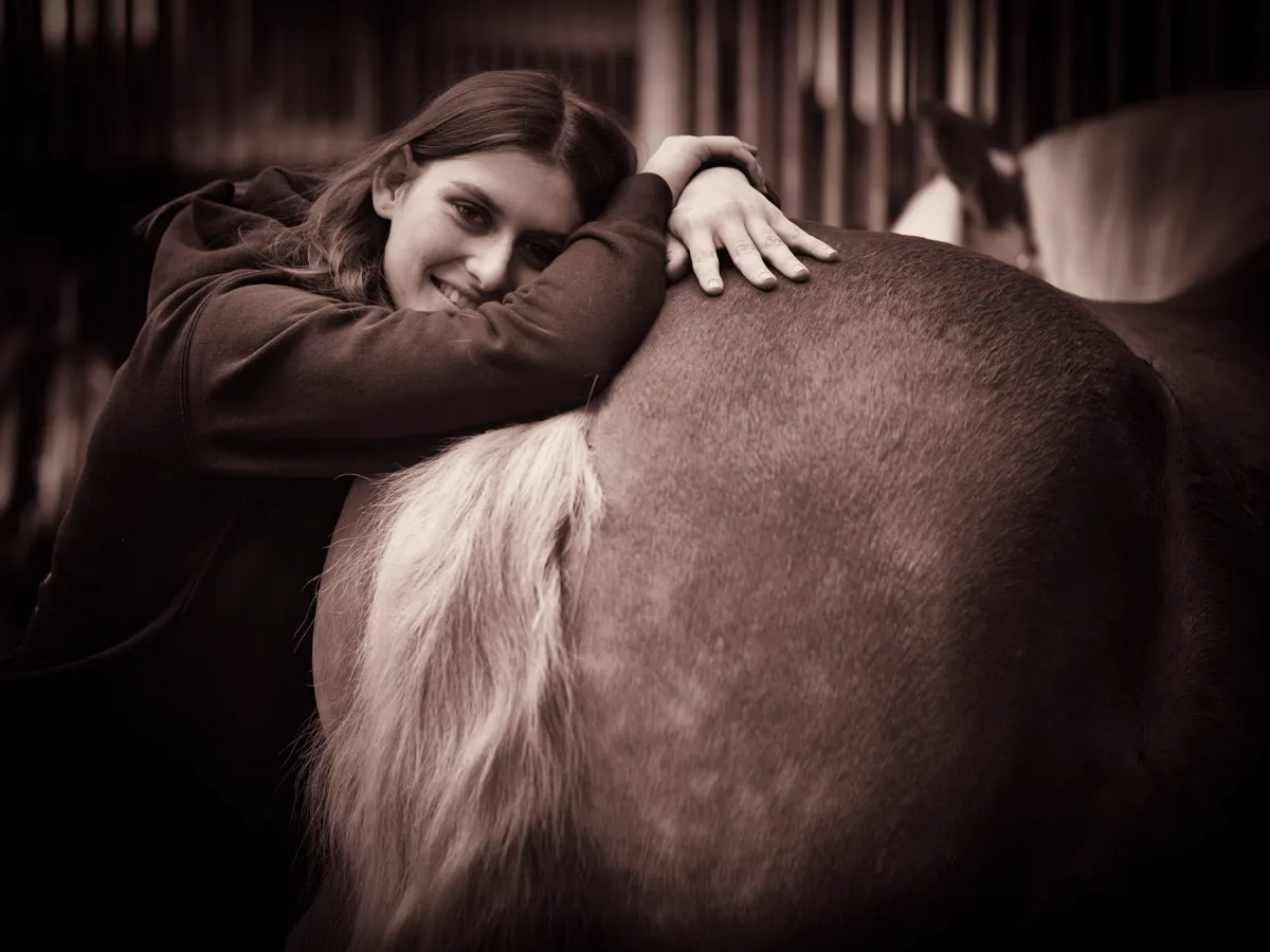 Lächelnde junge Frau lehnt sich kuschelnd an die Kruppe eines Pferdes mit heller Schweifrübe im Stall, Sepia