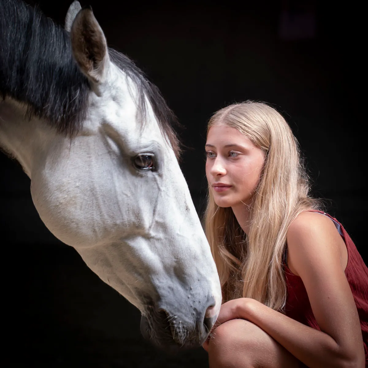 Blonde junge Frau hockt Gesicht an Gesicht mit einem Schimmel vor dunklem Hintergrund